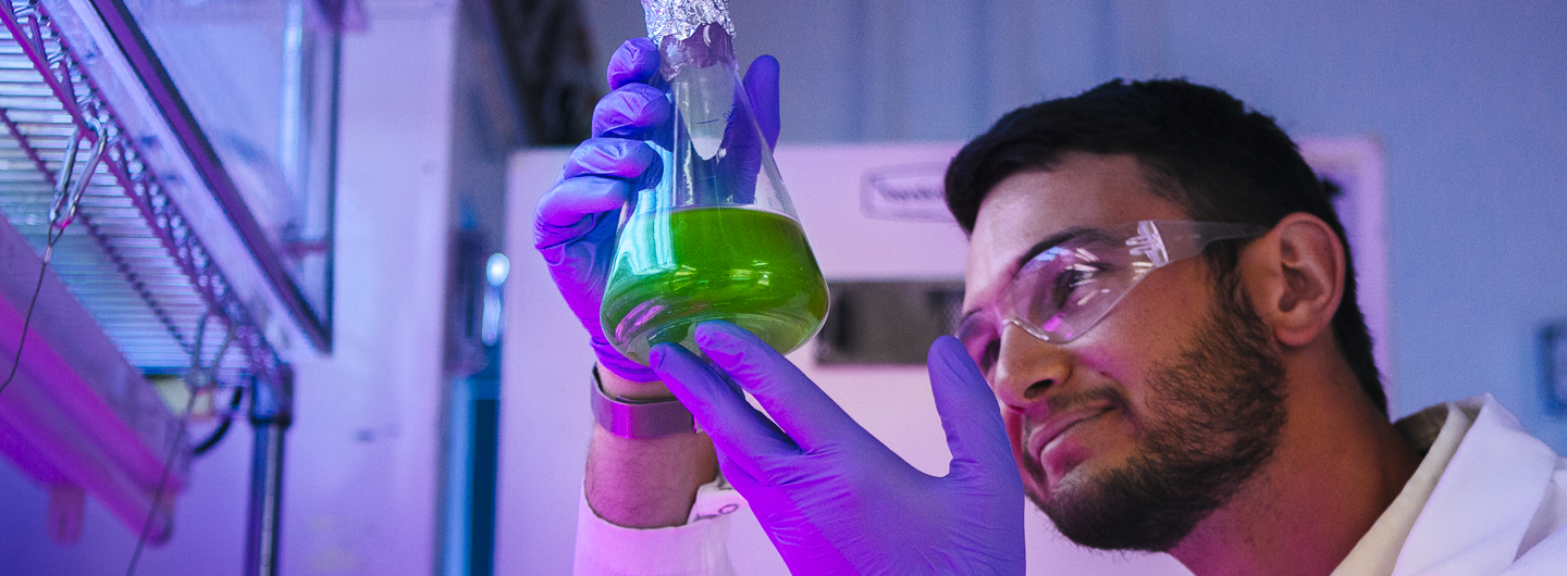 Male scientist in the lab with safety goggles on examining a flask with a green substance inside